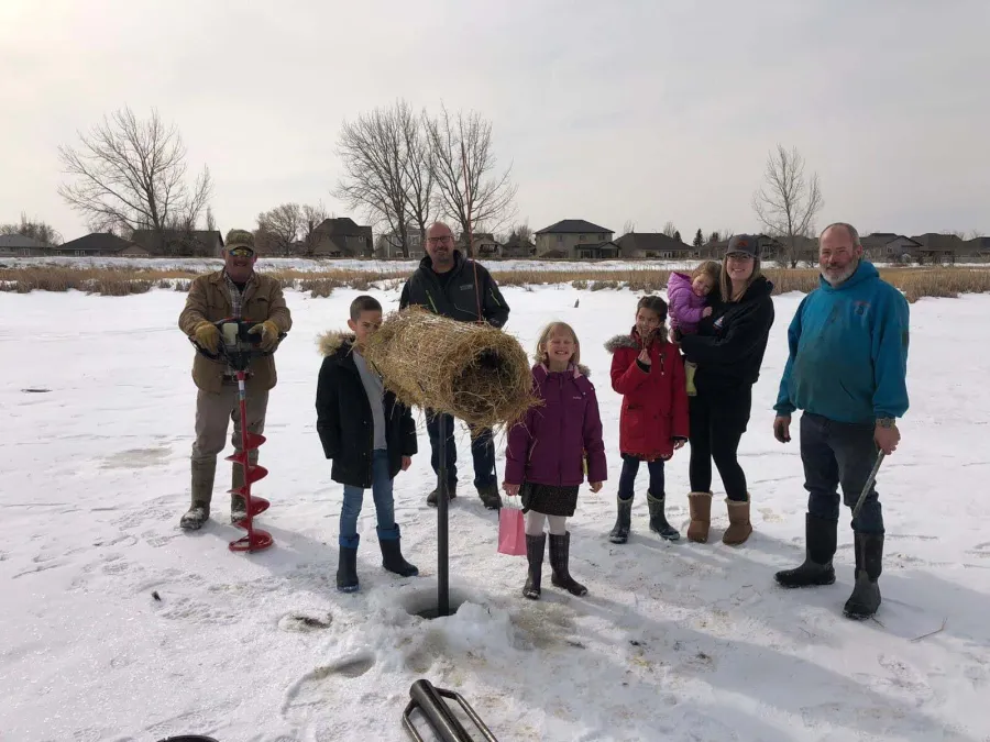Adults and kids on the ice with an ice auger, beside a straw-filled wire tube set on a post