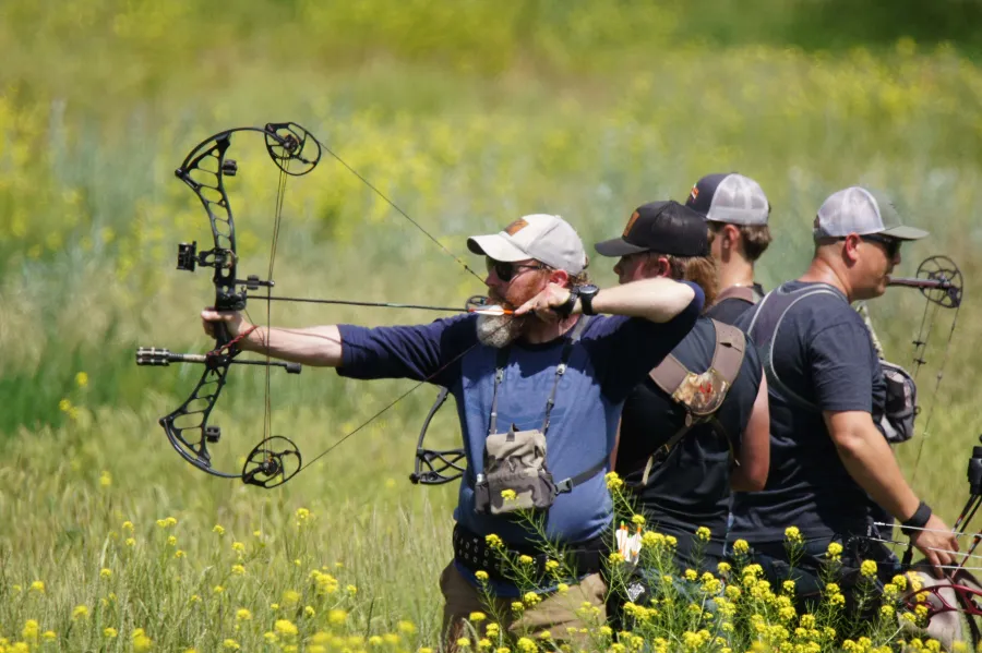 An archer at full draw with a compound bow, other archers lined up behind him in a field of yellow flowers